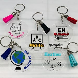 May include: Six clear acrylic keychains with colorful tassels. Each keychain features a different design, including a globe, a chalkboard, a book, a beaker, and math symbols. The keychains are personalized with names and titles, such as "Miss Lei", "Mr. Gauthier", and "SRA. VARELA".