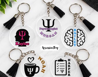 May include: Five circular clear acrylic keychains with silver metal rings and black tassels. Designs include a silhouette with a pink psi symbol and name Kaylee, a black psi symbol with text Psychologist Morgan, a brain with blue name Bailey, a coffee mug with name Noah, and a clipboard with name Harper.