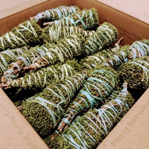 May include: A box filled with multiple bundles of dried green sage. Each bundle is wrapped with light blue and white twine. The sage bundles are arranged inside a brown cardboard box.