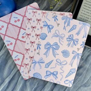 May include: Three decorative rectangular cards with pastel pink and white patterns. One card features a cherry grid design, another has red bows, and the third has blue bows, keys, and seashells. A blue candle and an e-reader are in the background.