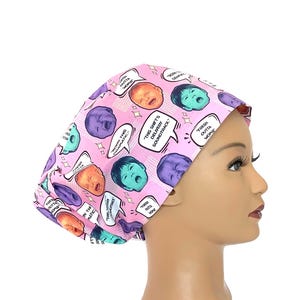 May include: A pink scrub cap featuring a repeating comic-style pattern of crying infants in various colors, including purple, teal, and orange. The design includes speech bubbles with the text "FRESH OUTTA WOMB," "BORN THIS DRAMATIC," and "THIS SHIFT'S DELIVERY SOUNDTRACK."
