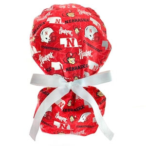 May include: A red scrub cap featuring a repeating pattern of white Nebraska Huskers logos, football helmets, and the word Nebraska. The cap includes a white ribbon tied in a bow at the back to secure the fabric, creating a gathered ponytail style.