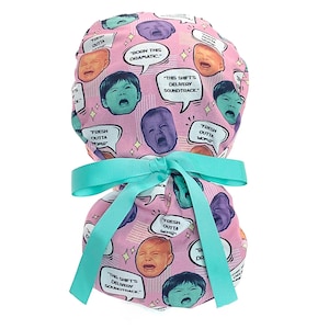 May include: A pink scrub cap featuring a repeating pattern of crying baby faces in various colors and speech bubbles with the text "BORN THIS DRAMATIC," "FRESH OUTTA WOMB," and "THIS SHIFT'S DELIVERY SOUNDTRACK." The cap is tied with a teal ribbon.