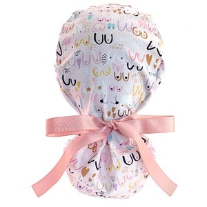 May include: A scrub cap featuring a light-colored fabric with a repeating pattern of stylized breast illustrations in black, pink, and gold, interspersed with small hearts, stars, and the words powerful and awesome, finished with a pink ribbon tie.