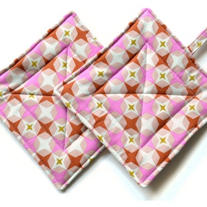 Puede incluir: Dos agarraderas de olla acolchadas con un patrón geométrico en tonos rosa, marrón y dorado. Las agarraderas son cuadradas y están hechas de tela.