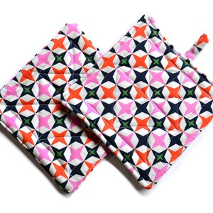 Puede incluir: Un par de agarraderas de olla acolchadas con un patrón geométrico de estrellas rosas, naranjas, verdes y negras sobre un fondo blanco.
