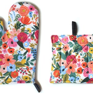 Puede incluir: Juego de manopla y agarradera con estampado floral. La tela blanca está cubierta con un colorido estampado floral con flores rosas, naranjas, azules y amarillas. La manopla tiene un lazo negro para colgar.