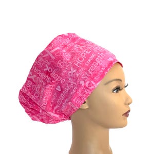 May include: A pink scrub cap featuring a repeating pattern of white awareness ribbons and inspirational words including HOPE, Strength, COURAGE, and survivor. The fabric has a mottled pink texture and is worn on a mannequin head.