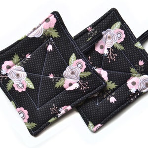 Puede incluir: Dos agarraderas de olla acolchadas negras con un patrón floral en rosa, blanco y verde. Las agarraderas tienen un fondo negro con lunares y un lazo negro para colgar.