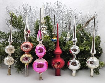 May include: A collection of vintage glass Christmas tree toppers in various colors, including silver, pink, red, and white. The toppers feature different designs, including a star, a butterfly, and a swirl.