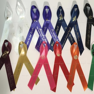 May include: A collection of colorful satin ribbons, each with personalized text and decorative elements. The ribbons vary in color, including white, gold, pink, red, and blue. Some ribbons feature text such as "Forever in our hearts" and names with dates.