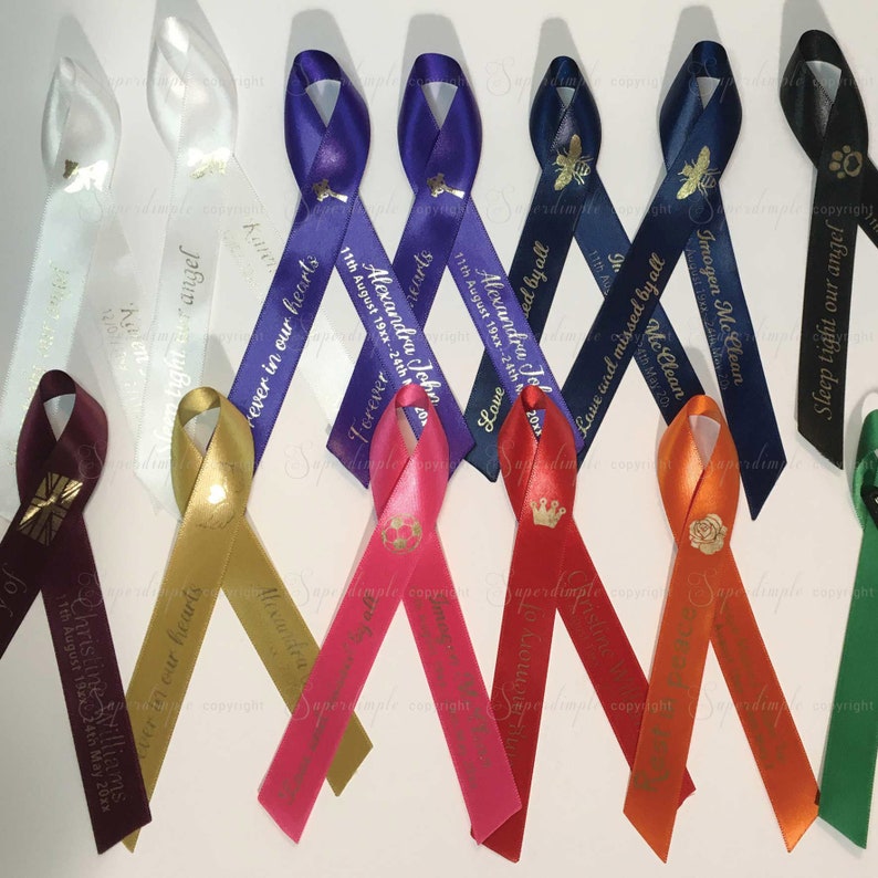 May include: A collection of colorful satin ribbons, each with personalized text and decorative elements. The ribbons vary in color, including white, gold, pink, red, and blue. Some ribbons feature text such as "Forever in our hearts" and names with dates.