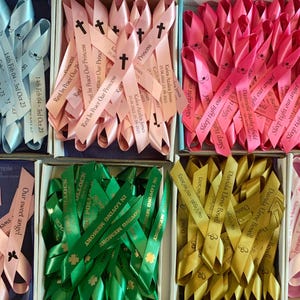 May include: Assortment of satin ribbons in various colors, including light blue, pink, green, and gold, each inscribed with personalized text and dates. The ribbons are arranged in boxes, with some featuring cross symbols and the words "Rest in Peace" and "In Loving Memory."