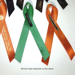 May include: Satin ribbon brooches in orange and green, with safety pin closures. One orange ribbon has text: "Joseph Mallord William Tur, August 19xx - 24th May 2." Additional black ribbons with the word "Sleep" are also visible.