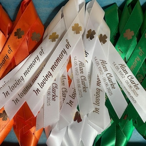 May include: A collection of orange, white, and green satin ribbons featuring the printed text In loving memory of, the name Alan Clarke, the dates 14.05.1952 - 09.06.2025, and a four-leaf clover icon. The ribbons are arranged in a layered overlapping pattern.