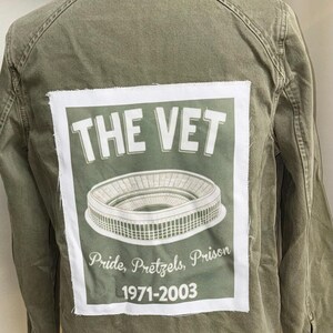 Op de afbeelding: Olijfgroene jas met een wit en groen grafisch ontwerp op de rug. De afbeelding toont "THE VET" boven een stadionillustratie, met "Pride, Pretzels, Prison 1971-2003" eronder. De jas is casual.