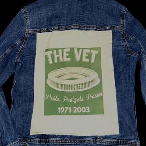 Op de afbeelding: Blauwe spijkerjas met een witte patch op de rug. De patch toont een groene stadionafbeelding met de tekst "THE VET" en "Pride, Pretzels, Prison 1971-2003". De jas heeft knopen en zakken.