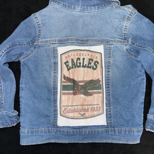 Könnte beinhalten: Hellblaue Jeansjacke mit einem Aufnäher mit dem Logo der Philadelphia Eagles. Der Aufnäher enthält den Teamnamen, eine Adlergrafik und den Text "Established 1933". Die Jacke hat einen klassischen Kragen und Knopfverschlüsse.