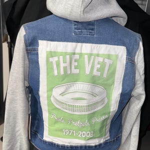 Op de afbeelding: Blauwe spijkerjas met grijze mouwen en een grijze capuchon. De achterkant heeft een witte rechthoek met een groene achtergrond, de woorden "THE VET", een stadionillustratie en de tekst "Pride, Pretzels, Prison 1971-2003."