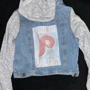 Peut inclure: Veste en jean avec un sweat à capuche gris attaché. Le dos de la veste présente un graphisme délavé du logo de l'équipe de baseball des Phillies de Philadelphie et le nom de l'équipe. La veste a deux boutons marron.