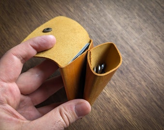 Könnte beinhalten: Eine braune Ledergeldbörse mit einer Klappe, die sich öffnet und einen Kartensteckplatz und ein separates Fach für Münzen freigibt. Die Geldbörse wird von einer Hand offen gehalten.