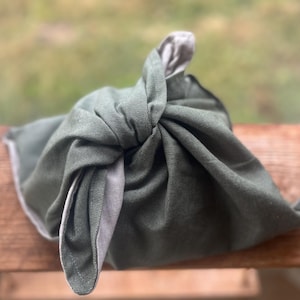 Puede incluir: Un envoltorio de tela verde atado con un nudo, con un interior gris visible. El envoltorio está hecho de un material texturizado y parece reutilizable. El fondo está borroso, lo que sugiere un entorno al aire libre. Este artículo probablemente se utiliza para envolver regalos o transportar objetos.