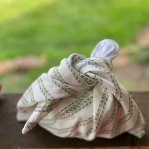 Peut inclure: Un paquet de tissu noué. Le tissu blanc est orné de rayures vert olive et d'un motif de feuilles répétitif. L'arrière-plan est un vert flou, suggérant un cadre extérieur.