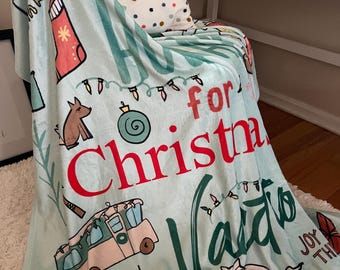 May include: A light blue Christmas-themed blanket with various illustrations, including a Christmas stocking, dog, ornament, and a camper van. The words "for Christmas" are in red, and "Vaca" is in green.