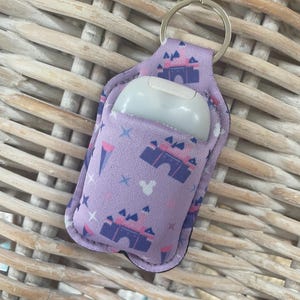 Könnte beinhalten: Lilafarbener Handdesinfektionsmittel-Halter mit einem Schloss- und Mickey-Mouse-Druck. Der Halter hat einen silbernen Schlüsselring und eine transparente Plastiktasche für die Desinfektionsmittelflasche.