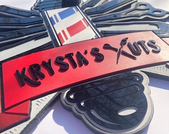May include: A layered sign with a barber shop theme. The sign features a red banner with the text "KRYSTA'S KUTS" in black, along with barber tools and a barber pole design. The sign is black, red, white, and gray.