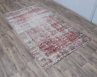 May include: Rectangular distressed rug with a faded red and cream color palette. The rug features a central floral pattern and a decorative border in shades of red, cream, and gray. The rug is placed on a wooden floor.