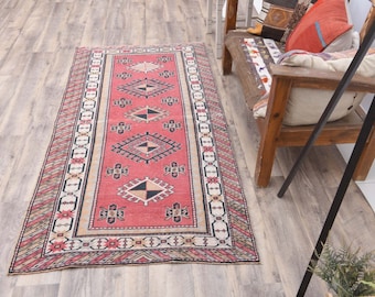Puede incluir: Una alfombra rectangular de estilo vintage con un campo central rojo. La alfombra presenta un patrón geométrico repetitivo en negro, blanco y beige, enmarcado por un borde con un diseño similar. La alfombra está colocada sobre un suelo de madera clara.
