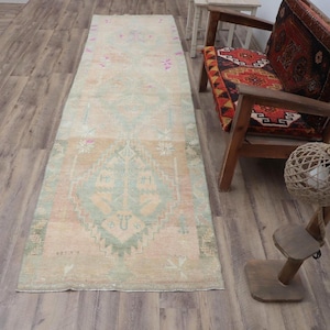 Op de afbeelding: Een rechthoekig vloerkleed in vintage-stijl met een vervaagde, verweerde uitstraling. Het vloerkleed heeft een centraal ontwerp in tinten beige, lichtblauw en perzik, met ingewikkelde geometrische patronen en bloemmotieven. Het vloerkleed ligt op een houten vloer.