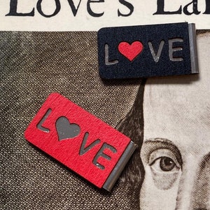 May include: Two black and red bookmark clips with the word "LOVE" and a heart in the centre. The bookmark clips are on a page of a book with the text "Love's Labours Lost" visible.
