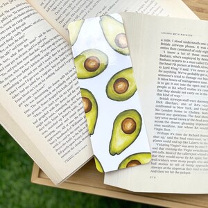May include: A bookmark featuring a repeating pattern of avocado halves with a white background, placed between the pages of an open book. The book's pages are visible, along with a wooden surface.