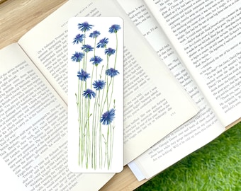 May include: A rectangular bookmark features a watercolor illustration of blue cornflowers on slender green stems. The floral design is set against a white background, with the flowers' delicate petals and green leaves adding a touch of nature to the reading accessory.