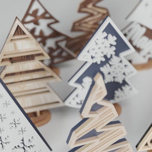 May include: Assortment of wooden Christmas tree ornaments in various styles and colors. Some trees are decorated with snowflakes, while others have a more rustic look.