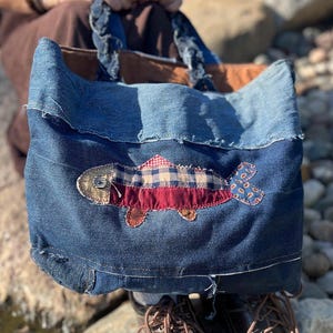 May include: A denim tote bag with a fish applique. The bag is made from various shades of blue denim and features a fish design in red, blue, and tan fabrics. The handles are made of twisted denim. Brown leather shoes are visible in the foreground.