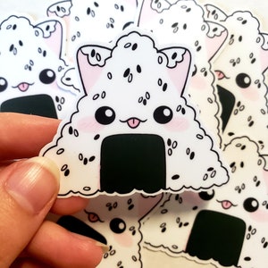May include: A collection of white, triangular stickers featuring a cute onigiri design with cat ears. The stickers have black sesame seeds, a black seaweed base, pink cheeks, and a smiling face with a tongue sticking out.