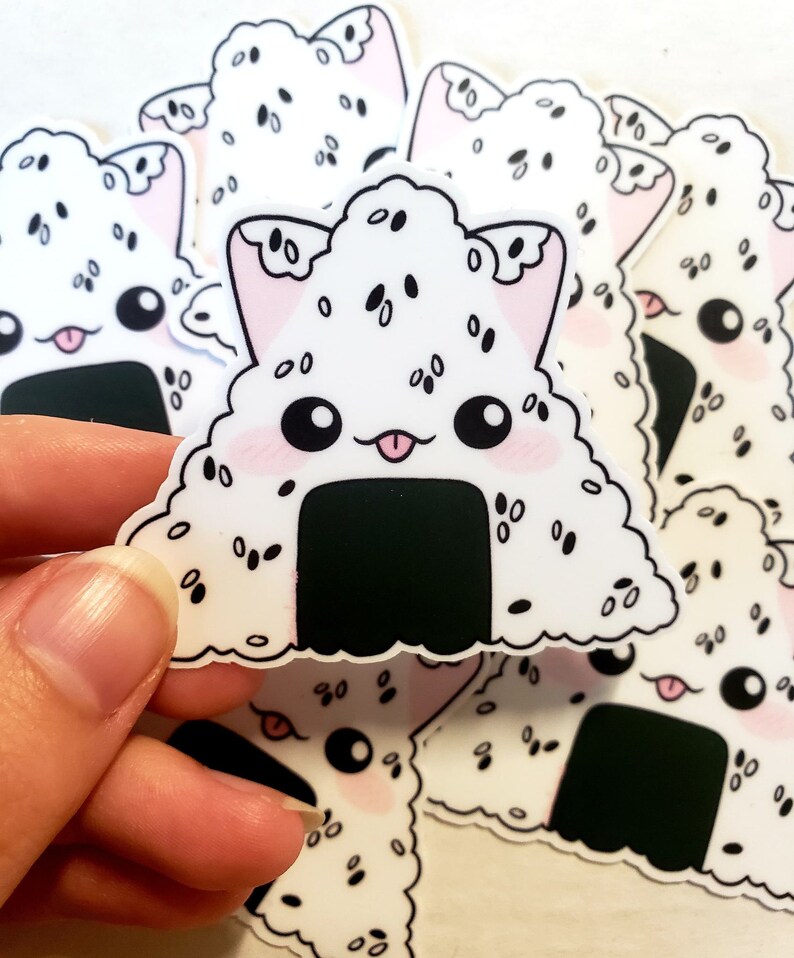 May include: A collection of white, triangular stickers featuring a cute onigiri design with cat ears. The stickers have black sesame seeds, a black seaweed base, pink cheeks, and a smiling face with a tongue sticking out.