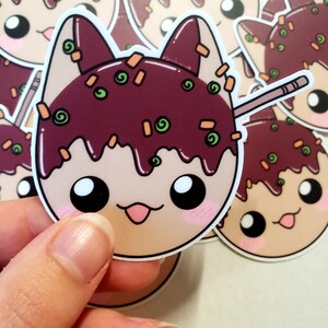 May include: A sticker featuring a cartoon cat head designed as takoyaki, a Japanese street food. The cat has a light brown face, dark brown ears, and a dark brown takoyaki topping with green and orange sprinkles. A wooden skewer is inserted into the takoyaki.