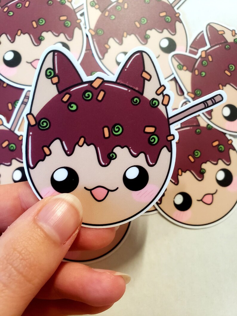 May include: A sticker featuring a cartoon cat head designed as takoyaki, a Japanese street food. The cat has a light brown face, dark brown ears, and a dark brown takoyaki topping with green and orange sprinkles. A wooden skewer is inserted into the takoyaki.