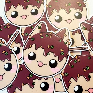 May include: A collection of stickers featuring a cartoon cat design. The cat's face is round with large eyes, a pink tongue, and blush. The cat's head is topped with a dark brown sauce and green and orange sprinkles. A wooden skewer is visible.