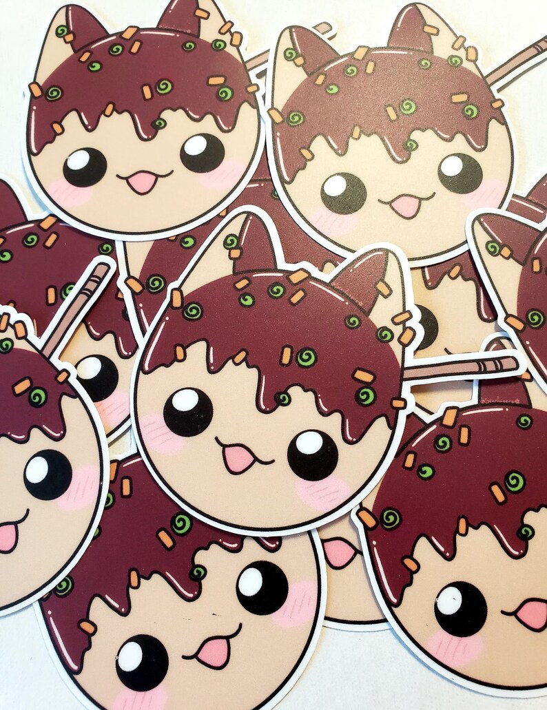 May include: A collection of stickers featuring a cartoon cat design. The cat's face is round with large eyes, a pink tongue, and blush. The cat's head is topped with a dark brown sauce and green and orange sprinkles. A wooden skewer is visible.