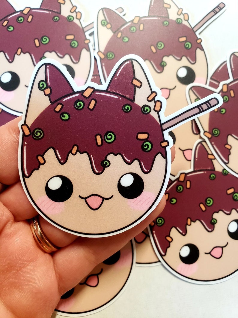May include: A collection of stickers featuring a cartoon cat design. The cat's head is styled to resemble takoyaki, with a dark brown topping, green and orange sprinkles, and a wooden skewer. The cat has large black eyes and a pink tongue.