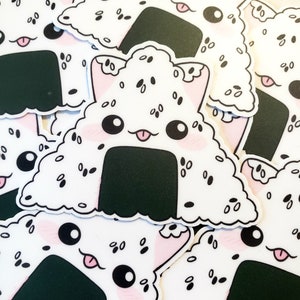 May include: A collection of white, triangular stickers featuring a cute onigiri design. The onigiri has a black seaweed wrap, pink cheeks, and a cat-like face with a tongue sticking out. The rice is speckled with black sesame seeds.