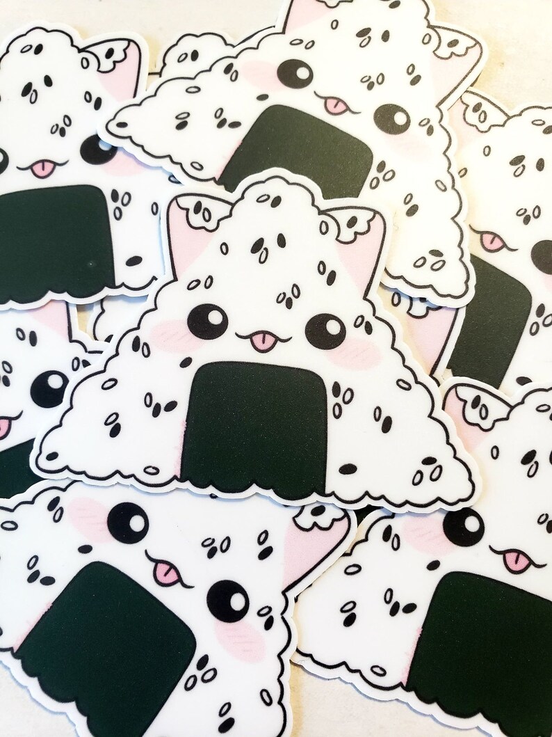May include: A collection of white, triangular stickers featuring a cute onigiri design. The onigiri has a black seaweed wrap, pink cheeks, and a cat-like face with a tongue sticking out. The rice is speckled with black sesame seeds.