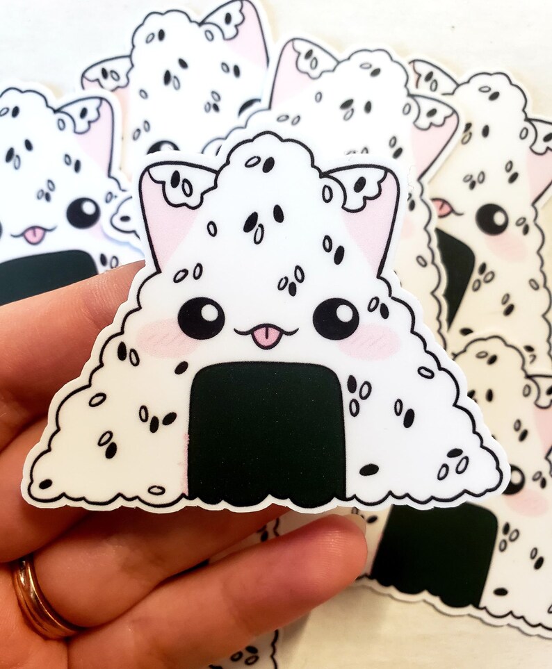 May include: A collection of white stickers featuring a cute onigiri design with cat ears. The onigiri has black sesame seeds, a black nori seaweed base, and a pink tongue. The stickers have a kawaii aesthetic.