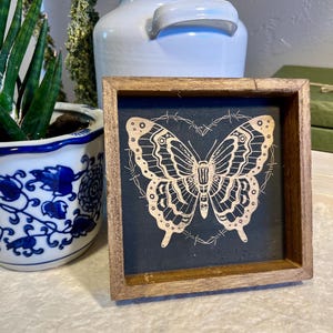 May include: A small, square wooden framed sign featuring a detailed butterfly design in a light color against a dark background. The butterfly is surrounded by a barbed wire design. A white ceramic jug and a potted plant are in the background.