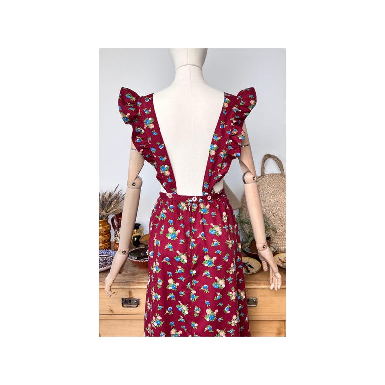May include: A burgundy sundress with a floral pattern of blue, white, and yellow flowers. The dress features ruffled straps and a low back. The dress is displayed on a mannequin.